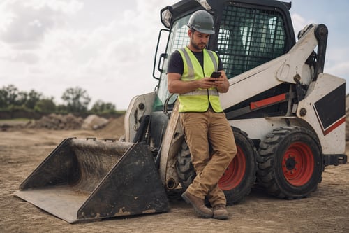 Compact Operator Reading Phone On Skid Steer Loader